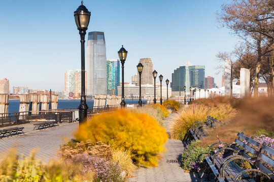 Waterfront walkway with park benches and lampposts overlooking the river and cityscape, representing urban travel and tourism in New York City, Manhattan, Jersey Skyline, USA.