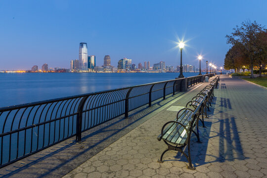 Empty park benches line a waterfront promenade at twilight, creating a peaceful scene for urban tourism and exploration with views of the Jersey City skyline, New York City, USA.