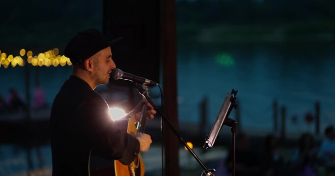Male guitarist singer performing on dimly lit outdoor waterfront stage at dusk