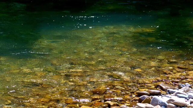 Crystal-clear river water flows gently over colorful stones in slow motion, revealing natural textures and reflections in stunning 4K detail.