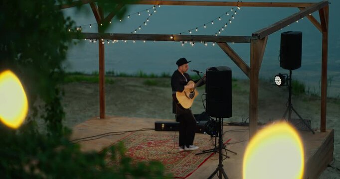 Solo guitarist performing on a small wooden waterfront stage under pergola lights