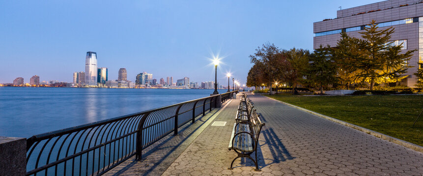 Benches line the Hudson River waterfront at dusk, providing a serene setting for urban travel and tourism with a view of the Jersey Skyline, Manhattan, New York City, United States.