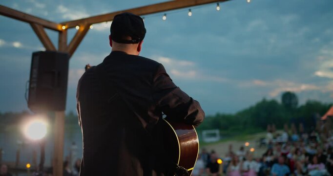 Rear view of guitarist performing on outdoor dusk stage with string lights