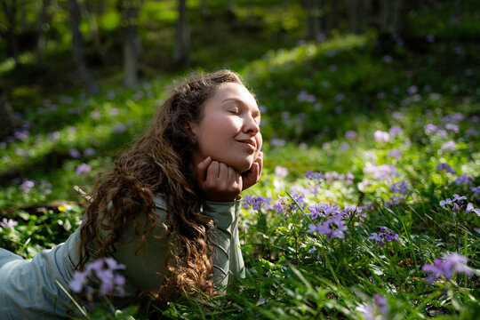 A woman lies in harmony on a green meadow, surrounded by wildflowers, with her eyes closed, hands under her chin. She is dreaming and taking a nap, enjoying the peaceful atmosphere of summer forest.
