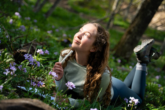 A woman in the summer park is taking notes in the sunlight, in a pensive mood thinking about the future. The girl has a diary where she plans goals and ideas for her creative lifestyle as a writer.