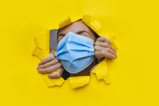 A woman wearing a protective medical mask that completely covers her face peeps through a torn hole in yellow paper.The concept of a pandemic,quarantine and protection against coronavirus and covid-19