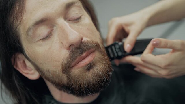 Professional female barber trimming beard of male client with electric clipper during grooming procedure in modern barbershop. Close up of hands and face.