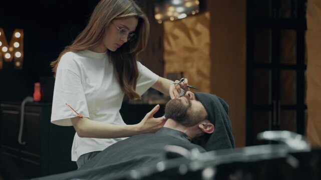Professional barber trimming beard of male client with scissors and comb during grooming procedure in modern barbershop salon