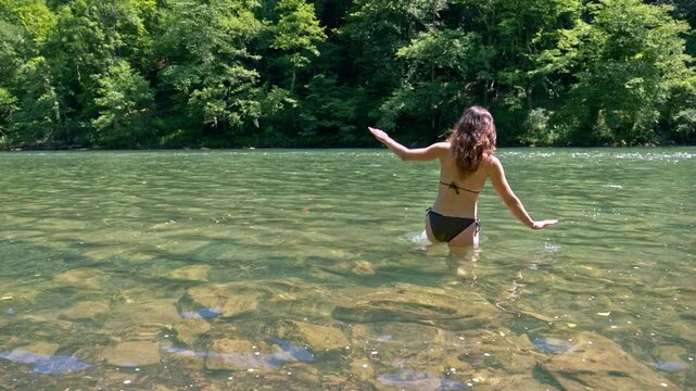 Woman enjoying a summer vacation in the crystal-clear waters of the Kolpa River, playfully wading through a pristine natural setting in slow motion 4K footage.