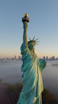 Slow vertical drone pan of Statue of Liberty at sunrise, New York. Low sea mist, clear sky. Manhattan skyline is visible. Copper patina illuminated monument.