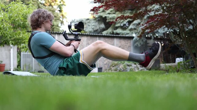 Young videographer performs a static core exercise while balancing with a gimbal stabilizer on grass improving endurance concentration and camera control