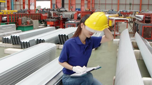 Female beautiful Asian worker with helmet and clipboard wiping sweat off forehead, working at industrial manufacturing factory. Tired staff inspect metal sheets. Inspector checking product quality.