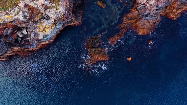 Rocky cliff edge meeting deep blue water, aerial perspective. Layered sedimentary rock with lichen and moss descending into teal sea, foam visible.