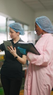 Quality Control in Action: Two food industry professionals, wearing hairnets and gloves, meticulously review product data on a tablet and clipboard in a sterile production environment.