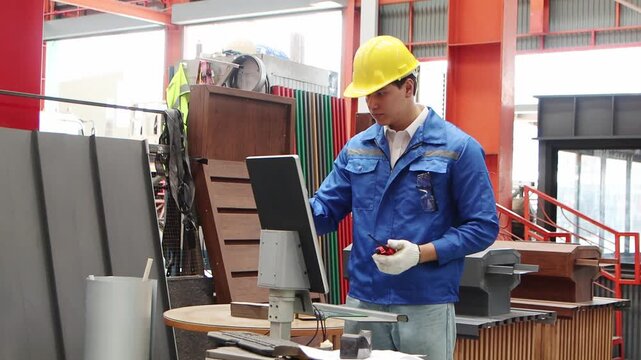 Young male worker with walkie talkie in safety clothing and helmet use desktop computer to operate machinery at manufacturing factory. Engineer working with industrial machine in industry site.