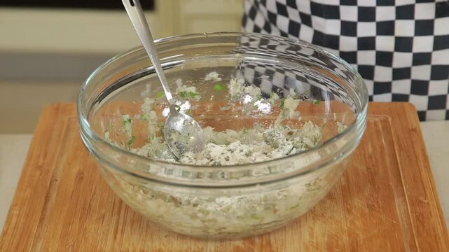 A chef mixing minced fish with egg and chopped fresh herbs in a glass bowl to prepare a mixture for fish patties