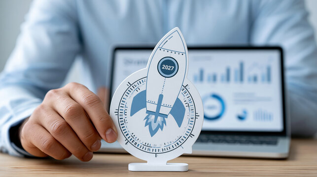 Rocket launch concept with 2027 year, business growth and success plan, hand holding paper rocket in front of laptop screen with charts
