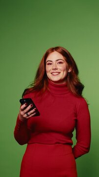 A young woman with long red hair, wearing a red turtleneck dress, smiles and interacts with her phone against a vibrant green screen background.