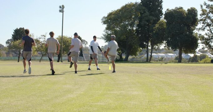 Performing warm-up drills diverse male athletes on grass field, with yellow cone, goal frame