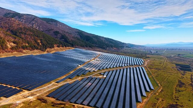 Drone perspective of solar farm between green hillside and village. Angled panel rows on sloped terrain with settlement and open fields in distance.