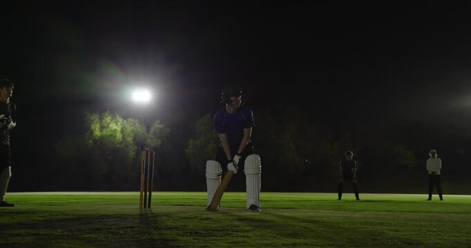 Male batsman reacting to run-up on cricket pitch at night, defending ball, getting out