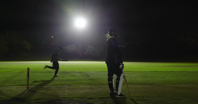 Three male friends playing floodlit cricket, delivering ball, crouching at stumps, copy space