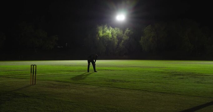 White male bowler running under lights delivering ball, fielder hitting stumps causing run-out