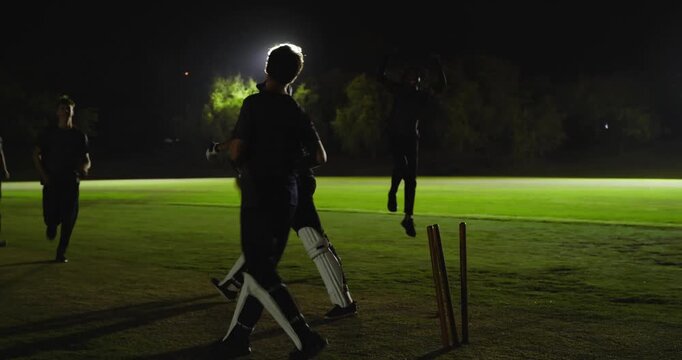 Diverse male cricket team reacting to bail drop, running and huddling, floodlights, cheering wicket