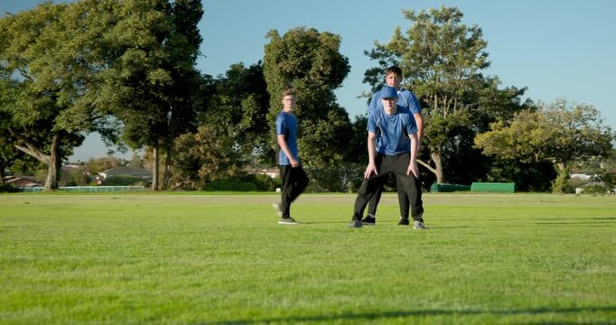 Diverse male cricket team on a field, bowler delivering ball, fielder lunging scooping