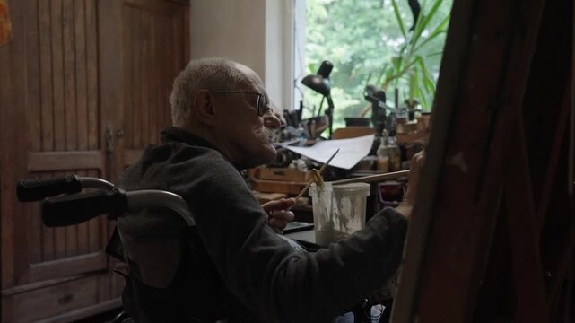 Senior artist on a wheelchair paints an oil painting on canvas in a studio