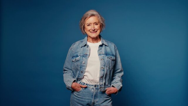 Smiling senior woman in denim jacket and jeans poses against a blue studio backdrop. She exudes confidence, style, and a positive attitude, capturing the essence of aging gracefully.
