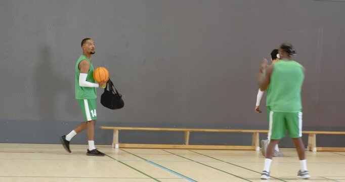 Diverse male teammates placing bags, drinking water and dribbling ball for warmup in basketball cour