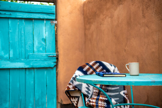 Cozy morning on a adobe patio in Santa Fe, New Mexico with a turquoise metal table with coffee, blanket and journal next to an adobe wall.