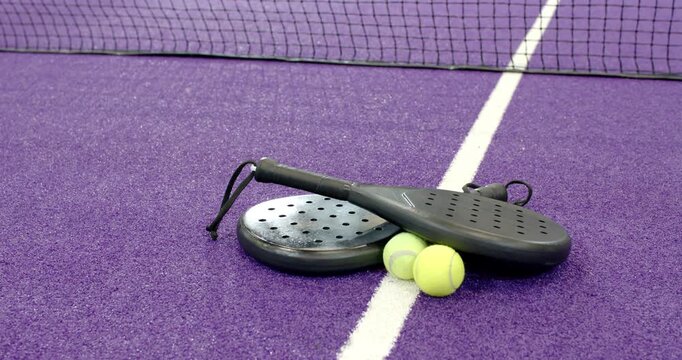 Camera starting overhead and capturing padel rackets and tennis balls resting by net on purple turf