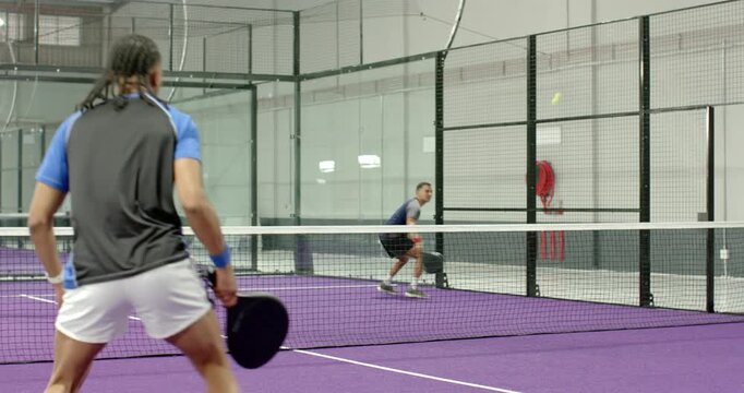 Lobbing ball over net, man with braided hair joining partner in rally on purple padel court