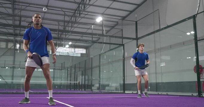 Diverse male athletes serving ball for practice, exchanging volleys, fist bumping on padel court