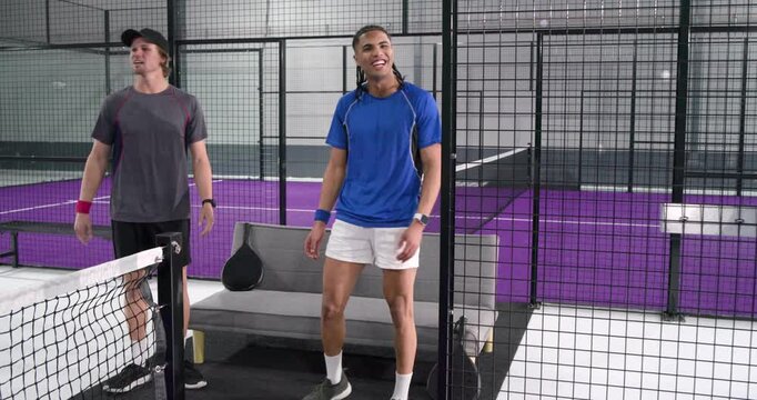 Male friends with rackets on padel court bench watching ball flying past net pumping arms