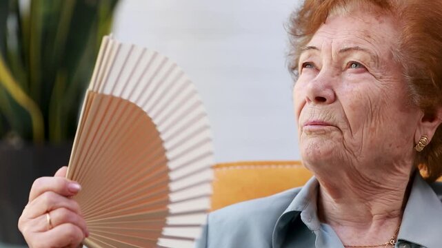portrait of a senior woman fanning herself with a fan while seated indoors; concept of summer heat wave and elderly people
