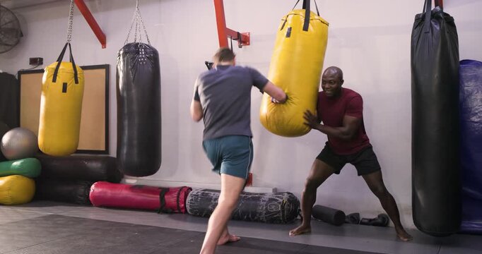 Male coach bracing yellow bag while male trainee alternating strikes, listening to him in gym