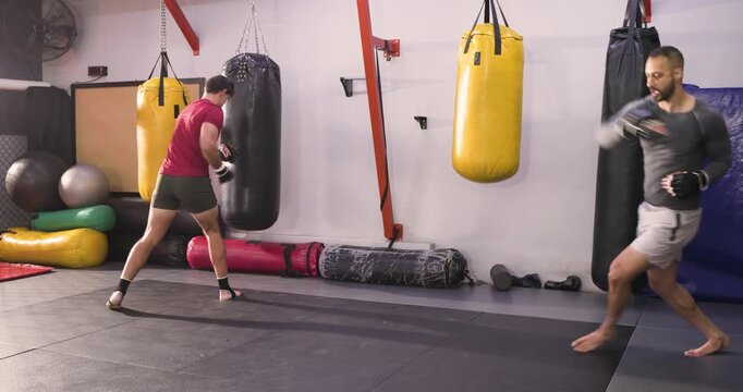 Diverse male training partners spotting heavy bags, executing punch and kick combos at dojo in gym