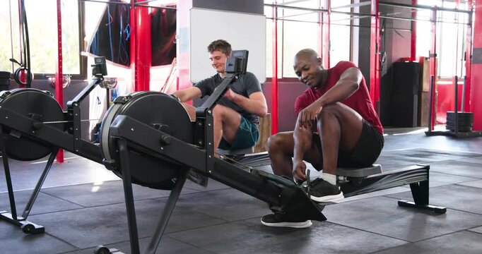 Exchanging smiles and sitting on rowing machines, diverse male friends rowing for workout