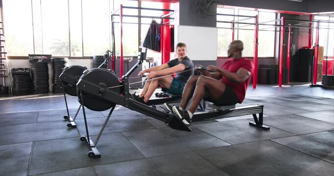 Diverse male friends rowing on gym machines, maintaining tempo for fitness