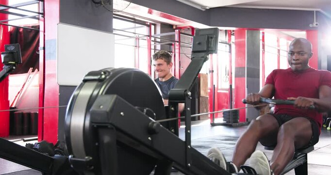 Diverse male friends rowing together on gym rowers after friend joining for fitness