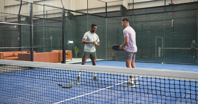 Meeting at net, diverse male friends volleying balls, practicing wrist flick on blue padel court