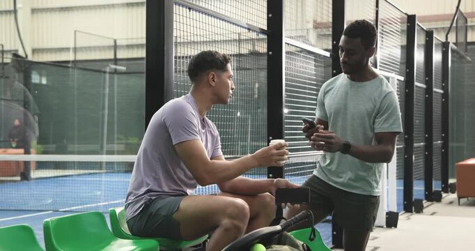 Diverse male friends sitting on bench at tennis courts, offering coffee and comparing phones