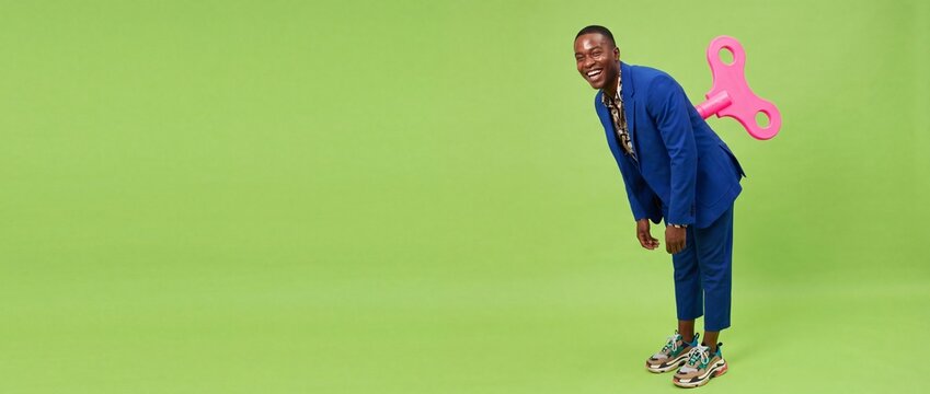 Happy Black man wind-up key in blue suit leaning forward on green background, representing corporate energy, automated motivation, and playful business concepts in a wide copy space banner format.