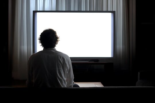 Man sitting on a couch watching a large blank television screen in a dark room with curtains