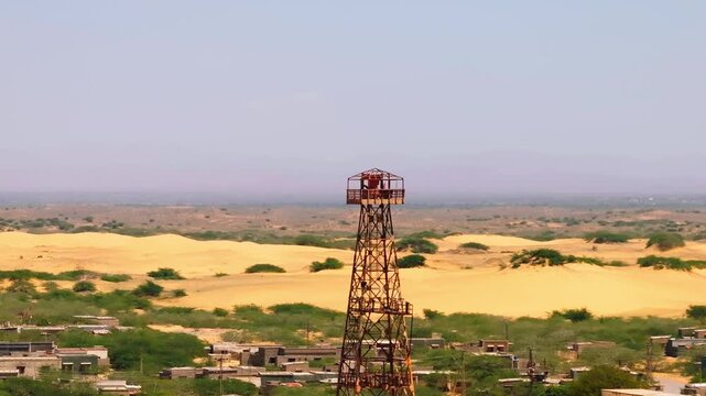 Large red steel tower framework on Damb Beach, with coastal houses and vast hazy plains behind. Downward tilting aerial ascent on hazy afternoon.