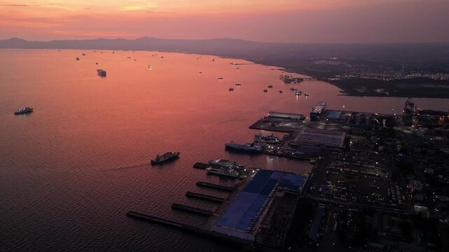Slow aerial shot of parked boats in Batangas Bay and one approaching baot at Batangas port in Batangas City Philippines with distant mountain silhouettes and nearby residential houses during sunset.