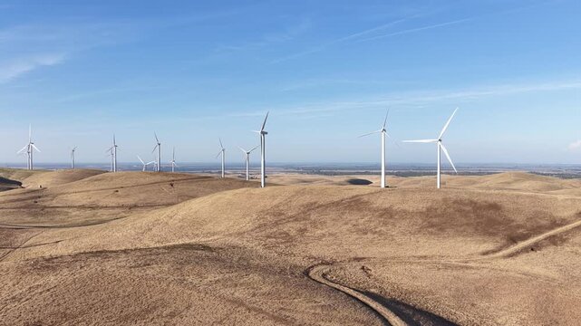 Drone footage of the Altamont Pass Wind Farm in California, showcasing numerous wind turbines spinning over rolling brown hills under a clear blue sky on a sunny day